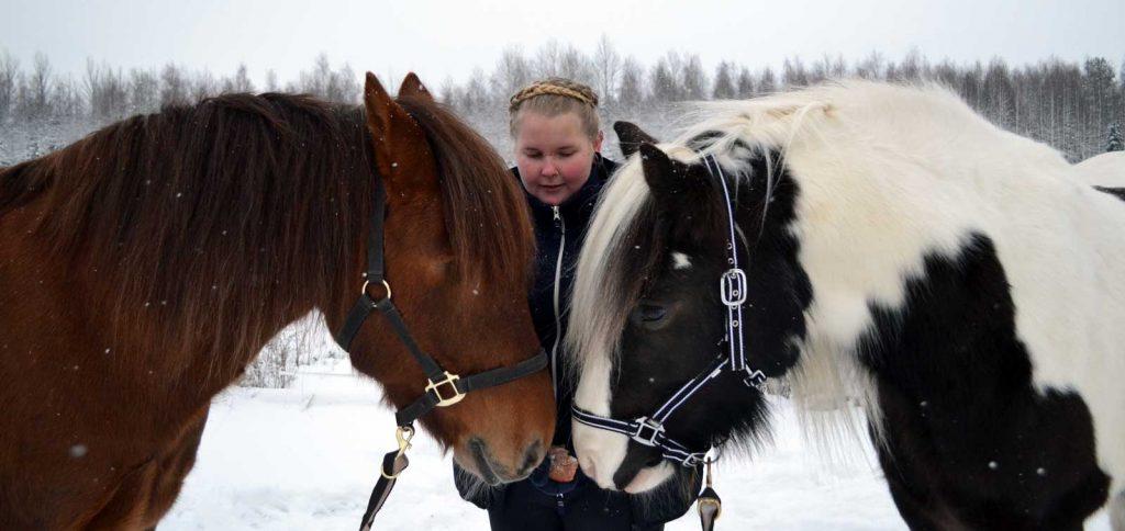 Kaksi hevosta turvat vastakkain, joiden takana blogin kirjoittaja Neea hevosia syöttämässä.