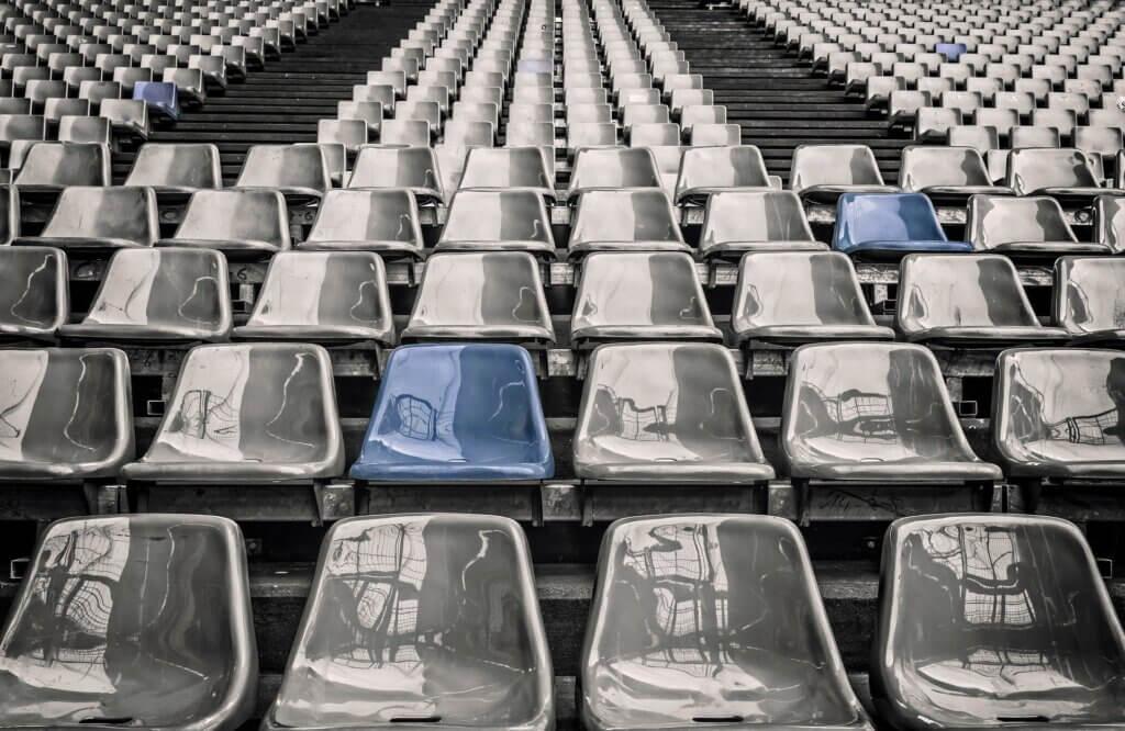Harmaasävyinen kuva suuresta stadionin katsomosta, jossa muutama tuoli on sinisen värisiä.