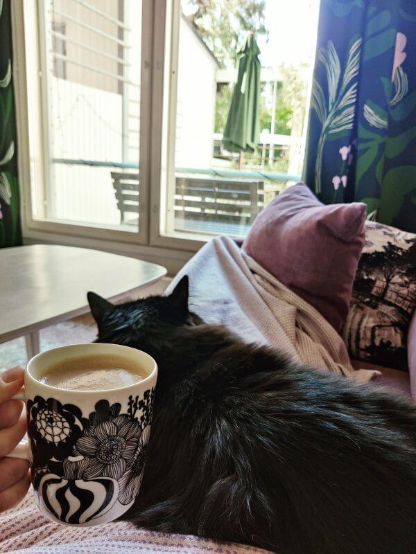 Kuvassa sohvalla pötköttelyä, sylissä kissa ja kädessä täysi kahvikuppi.